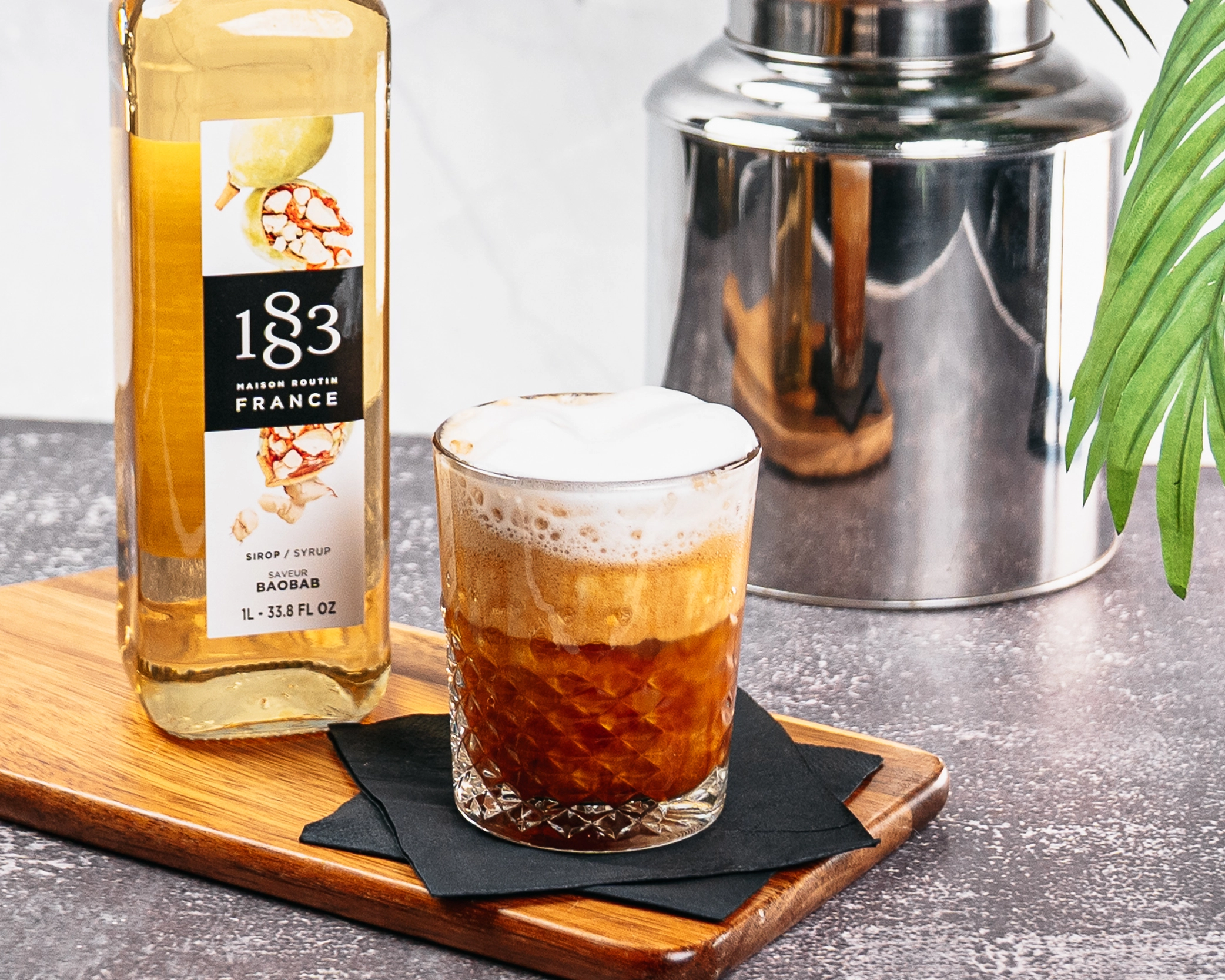 Espresso soda topped with cold foam beside a bottle of 1883 Baobab syrup on a wooden serving board.
